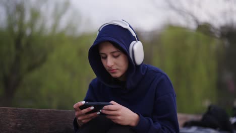 Mujer-Jugadora-Con-Capucha-Azul-Con-Auriculares-Usa-Smartphone,-Parque-Urbano-Al-Aire-Libre