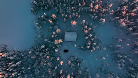 Cottage-in-the-snowy-woods,-winter-evening-in-Lapland---Birds-eye,-drone-shot