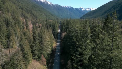 drone-shot-over-a-straight-road-following-a-driving-car-in-the-middle-of-a-forest-with-mountain-in-the-background,-chilliwack,-british-columbia,-canada