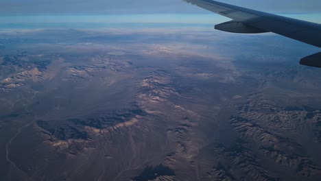 Flying-High-Above-Desert-Landscape-and-Barren-Fields-Passenger-POV-Over-Airplane-Wing