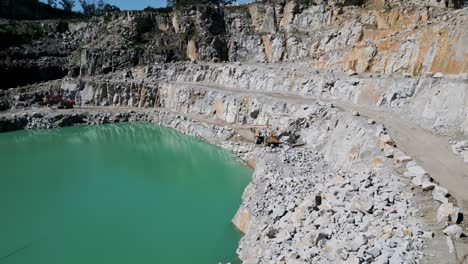 Excavator-loading-stone-into-a-truck-beside-a-turquoise-quarry-lake---aerial