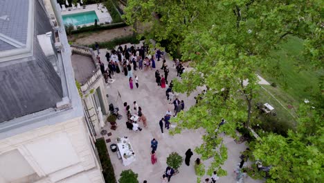 Luftaufnahme-Einer-Cocktailparty-Für-Eine-Hochzeit-Im-Freien-Auf-Der-Terrasse-Eines-Schlosses-Per-Drohne.-Links-Das-Schloss,-Rechts-Der-Garten-Und-Die-Bäume,-Im-Hintergrund-Ein-Swimmingpool,-Gäste-Beim-Spazierengehen