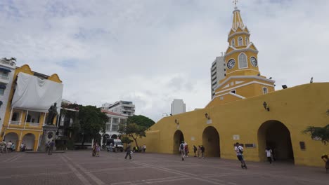 Gente-En-La-Plaza-Junto-Al-Monumento-Torre-Del-Reloj,-Fachada-Trasera-Amarilla-De-La-Torre-Del-Reloj,-Cartagena