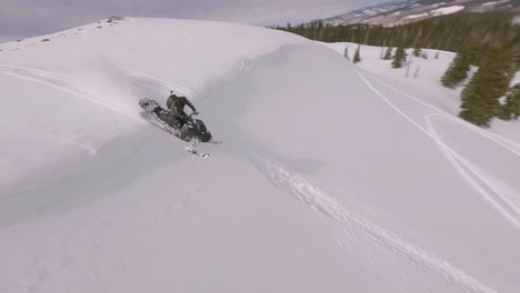 Luftaufnahmen-Von-FPV-über-Schneebedeckten-Hügeln-In-Der-Nähe-Des-Sharkstooth-Peak-Mit-Einem-Schneemobilfahrer,-Der-über-Die-Spitze-Fährt