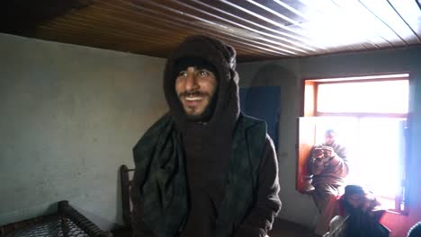 A-warmly-dressed-man-with-microcephaly-smiles-inside-a-home,-with-family-members-in-the-background