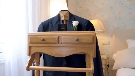 Muebles-De-Madera-Clara-Con-Las-Pertenencias-Del-Novio,-Su-Chaqueta-De-Traje-Con-Una-Rosa-Blanca-En-El-Bolsillo-Delantero,-El-Perfume-Colocado-En-Los-Muebles,-En-Una-Habitación-Muy-Luminosa-Con-La-Ventana-Y-Una-Cortina-Blanca