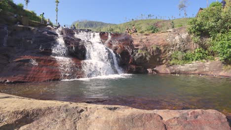 Breathtaking-drone-shot-of-Gartmore-Falls-in-Sri-Lanka,-featuring-lush-greenery,-orange-toned-sands,-and-rocks-against-a-vivid-blue-sky