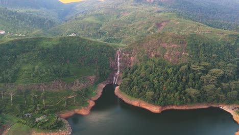 Breathtaking-drone-shot-of-Gartmore-Falls-in-Sri-Lanka,-featuring-lush-greenery,-orange-toned-sands,-and-rocks-against-a-vivid-blue-sky