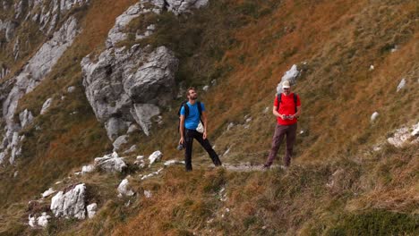 Aerial-View-Of-Two-Male-Hiking-Friends-On-Ridgeline-Filming-Themselves-Using-Drone-In-Lecco-Alps