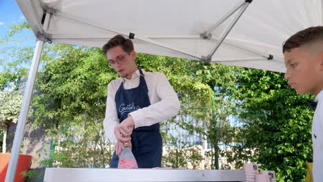 a-young-Caucasian-waiter-in-a-long-sleeved-white-shirt-and-dark-blue-apron-making-ice-creams-at-a-stand-with-a-spatula,-a-new-iceroll-concept-on-a-refrigerated-plate