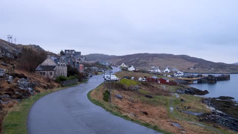 Solo-blue-car-driving-along-scenic-coastal-road-in-remote-Castlebay-village-on-the-island-of-Barra-in-the-Outer-Hebrides,-Scotland-UK