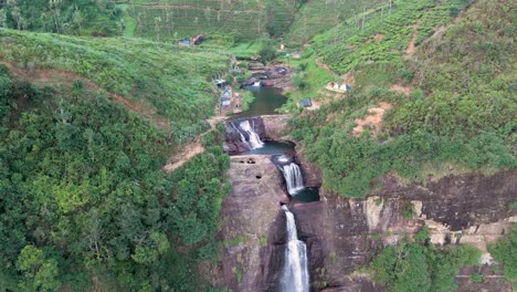 Breathtaking-drone-shot-of-Gartmore-Falls-in-Sri-Lanka,-featuring-lush-greenery,-orange-toned-sands,-and-rocks-against-a-vivid-blue-sky