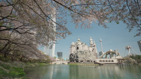 POV-Standing-Under-Cherry-Blossom-Trees-by-Seokchon-Lake-with-View-of-Lotte-World-Amusement-Park-and-Lotte-Tower-in-Spring-Seoul,-South-Korea---pan-right