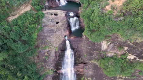 Atemberaubende-Drohnenaufnahme-Der-Gartmore-Falls-In-Sri-Lanka,-Mit-üppigem-Grün,-Orangefarbenem-Sand-Und-Felsen-Vor-Einem-Strahlend-Blauen-Himmel