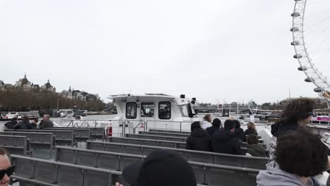 Passengers-enjoy-a-Thames-river-cruise-with-London-Eye-backdrop,-overcast-sky