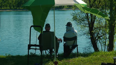 Una-Pareja-De-Ancianos-Disfruta-De-Un-Tranquilo-Día-De-Pesca-Bajo-Unas-Sombrillas-Junto-A-Las-Serenas-Aguas-Del-Lago-Jarun.