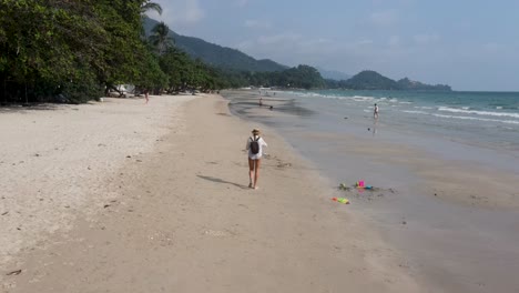 Low-Flying-Along-White-Sand-Beach-At-Koh-Chang-Following-Female-Beachgoer-Wearing-Backpack