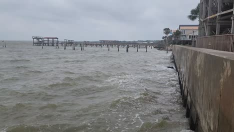 Dies-Ist-Ein-Kurzer-Videoclip-Von-Meereswellen,-Die-Während-Eines-Nachmittagssturms-Auf-Einen-Betonpier-Mit-Holzstegen-Und-Häusern-Im-Hintergrund-Krachen