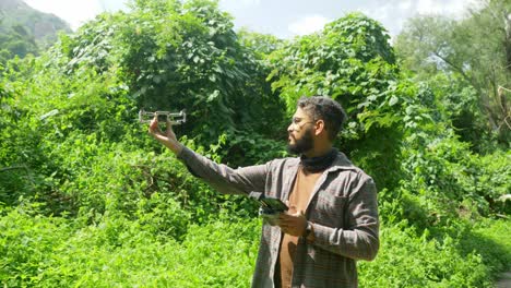 A-young-Indian-man-landing-his-mini-drone-in-a-wild-forest-landscape-for-wildlife-surveillance,-Western-Ghats,-India