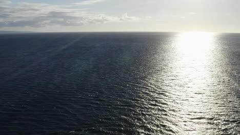 Aerial-dolly-out-to-sea-as-sunlight-ray-bounces-on-ocean-surface-with-ripples-and-current