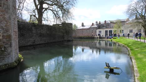 People-visiting-and-walking-around-the-historic-moat-of-the-Bishop's-Palace-grounds-in-city-of-Wells,-Somerset,-England