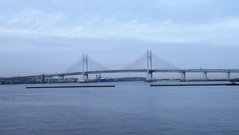 Wide-shot-of-Yokohama-Bay-Bridge-over-calm-water-with-cloudy-skies,-industrial-backdrop,-serene