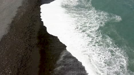 Waves-gently-washing-over-a-black-sand-beach,-aerial-view