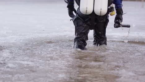 Buceador-De-Hielo-Equipado-Con-Traje-Seco-Camina-Hacia-El-Agua-Y-Se-Sienta-En-El-Borde-Del-Hielo-Del-Lago