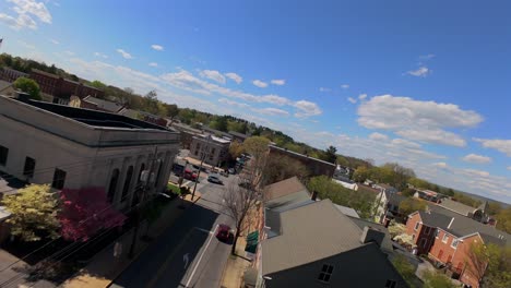 Small-town-street,-lined-with-red-brick-buildings-and-parked-cars,-under-a-clear-blue-sky-dotted-with-clouds