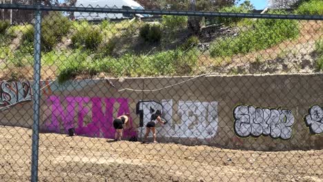 Two-female-taggers-illegally-spraying-graffiti-paint,-defacing-wall-in-the-Los-Angeles-area