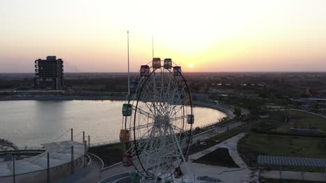 The-drone-camera-is-panning-to-the-right-where-the-sun-is-setting-between-the-giant-wheel