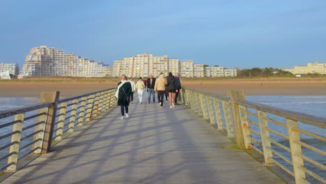 Saint-Jean-de-Monts,-France:-Shot-of-people-walking-on-wooden-pier-on-french-atlantic-coast-with-city-buildings-skyline-in-the-background-at-dusk