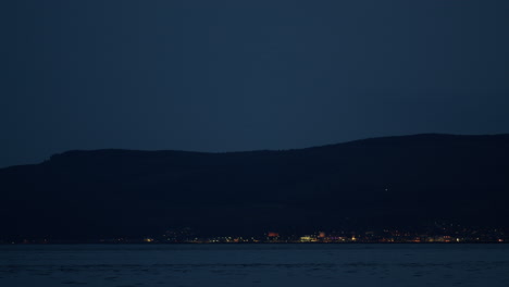 Water-ripples-pulse-across-surface-on-coast-with-lights-on-houses-under-mountains