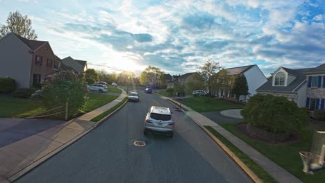 Drone-following-shot-of-silver-car-on-road-in-american-residential-area-during-sunset-time