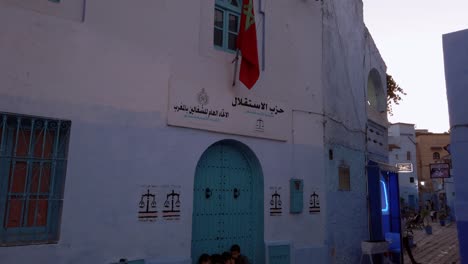 Un-Grupo-De-Niños-Mira-Un-Teléfono-Sentado-En-Una-Puerta-De-Entrada-De-Color-Bajo-La-Bandera-Roja-Marroquí-En-El-Centro-Histórico-De-Chefchaouen,-Marruecos