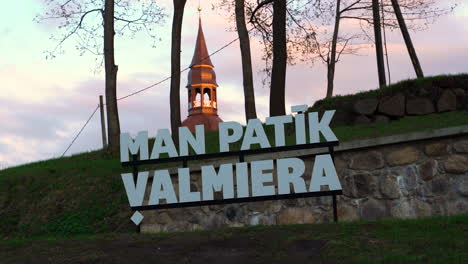 &quot;Ich-Mag-Valmiera&quot;-Schild-Mit-St.-Simons-Kirchturm-Im-Hintergrund-Mit-Sonnenuntergangshimmel