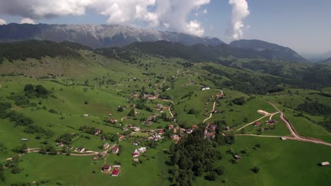 Sirnea-village-nestled-in-green-hills-with-church-and-mountains-in-the-backdrop,-sunny-day,-aerial-view