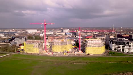 Backwards-aerial-showing-red-cranes-rising-above-construction-site-Noorderhaven-part-of-urban-development-and-housing-neighbourhood-seen-from-river-IJssel-floodplains