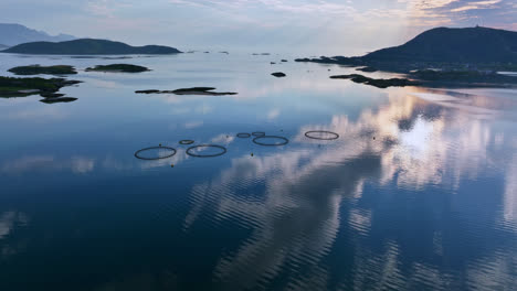 Drone-shot-circling-fish-cages,-sky-mirroring-from-the-water-surface,-in-Norway