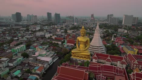 A-giant-golden-buddha-statue-in-urban-bangkok-at-dusk,-serene-atmosphere,-aerial-view