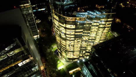 Aerial-view-establishing-an-office-building-illuminated-at-night,-modern-glass-building,-Nueva-Las-Condes-Santiago-Chile