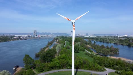 Wind-turbine-spinning-on-Danube-Island-with-Vienna-skyline-and-river,-sunny-day,-renewable-energy-concept,-aerial-view