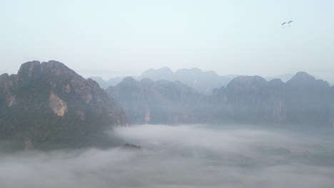 drone-shot-of-morning-fog-shrouding-cliffs-in-Vang-Vieng,-the-adventure-capital-of-Laos
