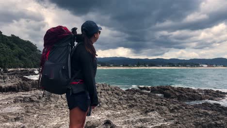 Female-with-sunglasses-and-hat-points-at-dark-clouds-in-the-distance-while-standing-on-rocky-ocean-shore