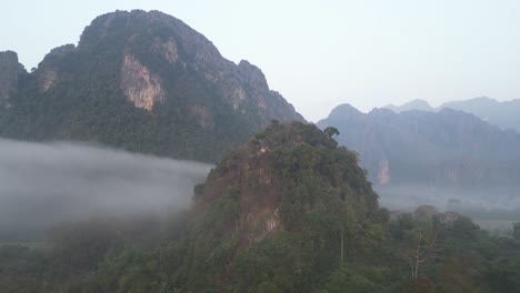 drone-shot-of-fog-rising-above-jagged-cliffs-in-Vang-Vieng,-the-adventure-capital-of-Laos