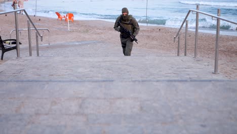 Ein-Bewaffneter-Soldat-Rennt-Die-Stufen-Vom-Strand-Hinauf,-Während-Ein-Paar-Am-Wasser-Entlang-Geht