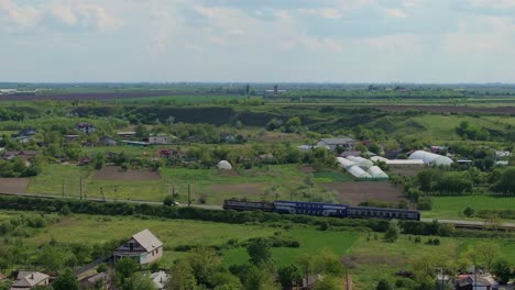 Rural-commute-in-an-old-blue-train,-travelling-between-houses-and-green-fields-on-a-sunny-clear-day,-aerial-tracking-shot