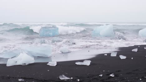 Wellen-Brechen-über-Eis-Und-Eisberge-An-Einem-Schwarzen-Sandstrand-In-Island---Zeitlupe