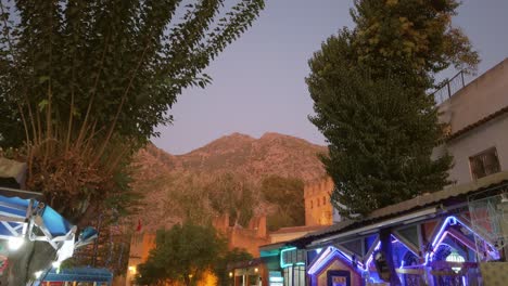 Vibrant-and-illuminated-town-centre-of-Chefchaouen-at-dusk,-with-tourists,-local-restaurants,-and-the-Rif-Mountains-in-the-background,-Morocco