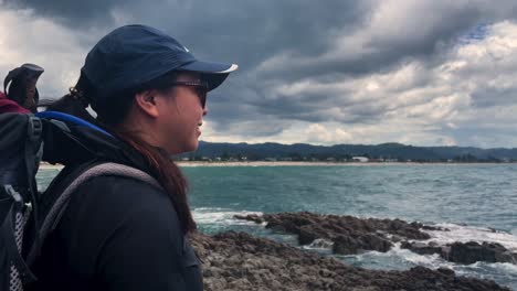 Young-asian-woman-with-backpack-smiles-as-she-looks-out-at-storm-above-the-ocean,-New-Zealand